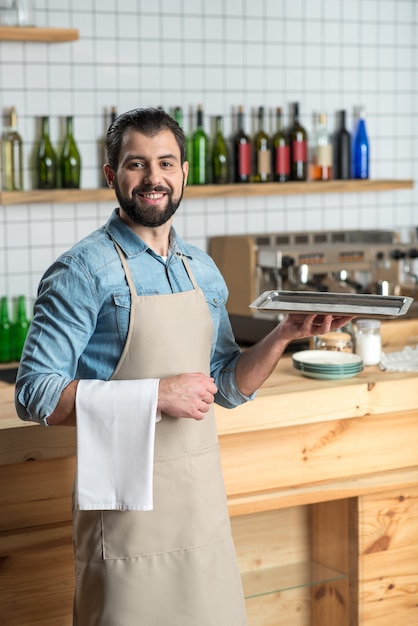 Foto camarero alegre. camarero sonriente barbudo experto que parece seguro mientras está en el trabajo y está listo para llevar el pedido con una bandeja en la mano