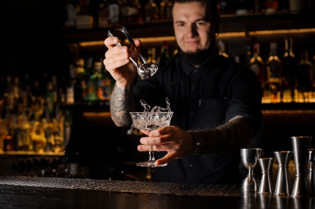 Camarero añadiendo a una bebida alcohólica en el vaso un gran cubo de hielo con pinzas en la barra del bar