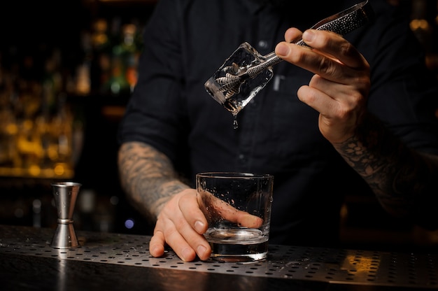 Camarero añadiendo a una bebida alcohólica en el vaso un gran cubo de hielo con pinzas en la barra del bar