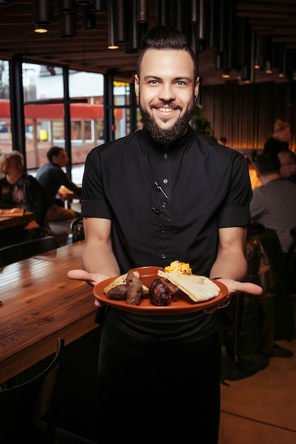 Camarero barbudo alegre con kebab, barbacoa y guarnición en un restaurante georgiano. Un camarero exigente vestido de negro, con barba y una ración de kebabs.