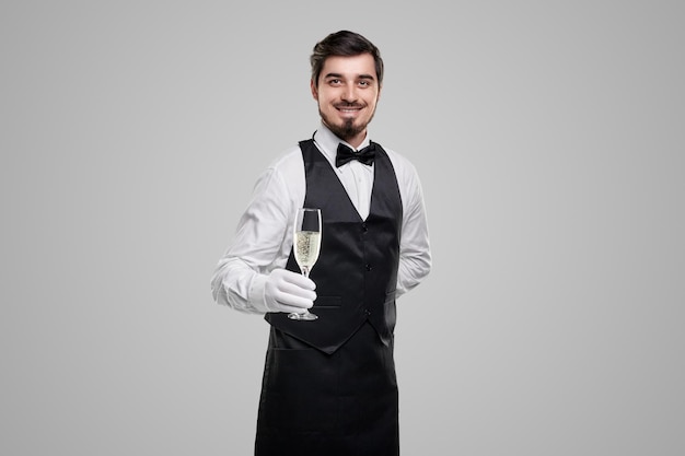 Camarero barbudo joven hermoso en uniforme elegante que ofrece una copa de champán y mirando a la cámara contra el fondo gris