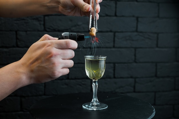 El camarero enciende el quemador sobre un delicioso cóctel alcohólico en una copa vintage para servir a los invitados. Bebida alcohólica caliente ardiente. De cerca