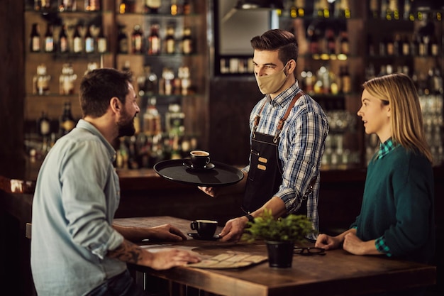 Camarero feliz con mascarilla mientras sirve café a una pareja en la cafetería