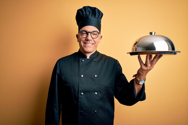 Camarero guapo de mediana edad con uniforme de cocina y sombrero sosteniendo una bandeja con una sonrisa feliz y fresca en la cara Persona afortunada