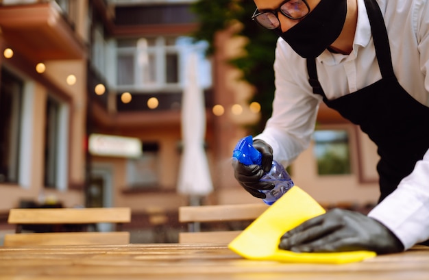 Camarero con máscara protectora y guantes desinfectando la mesa del restaurante para el próximo cliente.