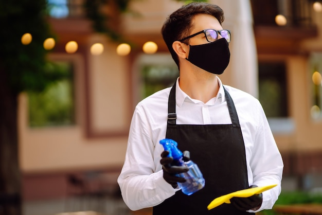 Camarero con máscara protectora y guantes desinfectando la mesa del restaurante para el próximo cliente.