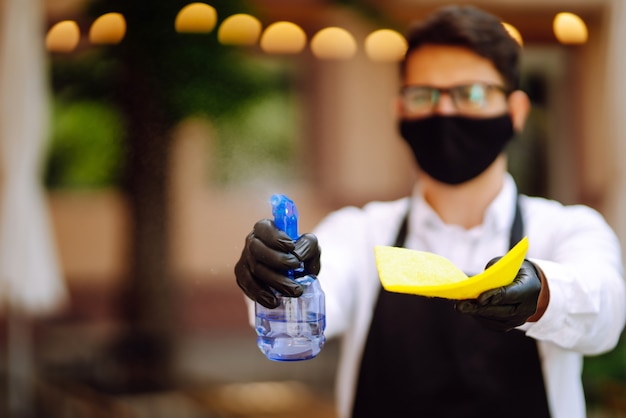 Foto camarero con máscara protectora y guantes desinfectando la mesa del restaurante para el próximo cliente.