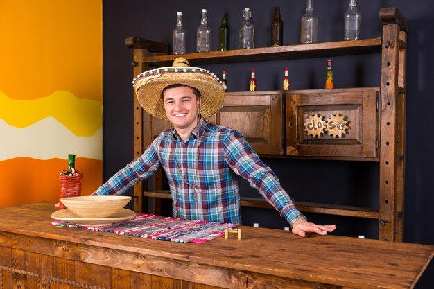 Camarero masculino guapo sonriente en un sombrero de pie en el mostrador en un pub mexicano, un estante con salsas picantes en el fondo