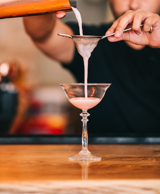 Camarero masculino vertiendo un delicioso cóctel de la coctelera de acero a través del colador a un vaso en la barra del bar en el oscuro fondo borroso