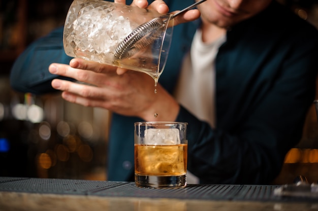 Camarero moreno vertiendo una bebida alcohólica en un vaso con un cubito de hielo