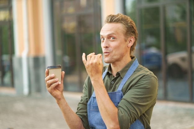 Camarero presentando una deliciosa y sabrosa taza de café en un vaso de papel para llevar con una sonrisa en la cara del camarero