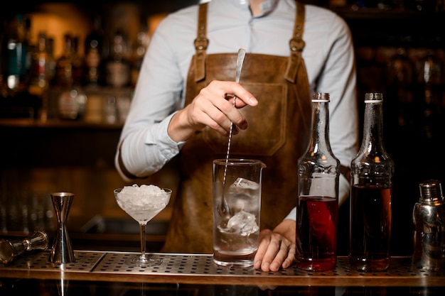 Camarero profesional masculino revolviendo una bebida alcohólica con hielo en la taza de vidrio de medición