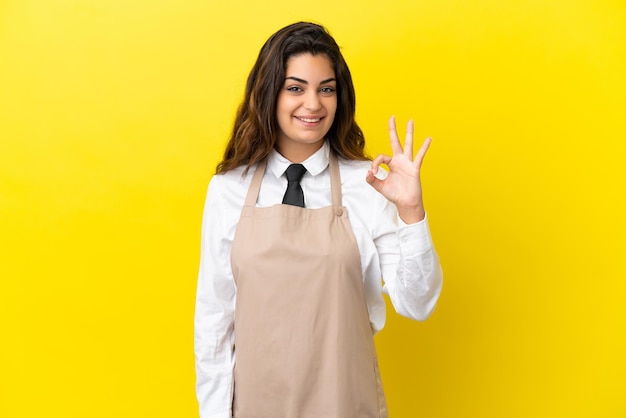 Camarero de restaurante caucásico joven aislado sobre fondo amarillo mostrando signo ok con los dedos