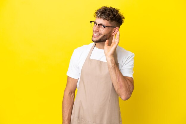 Camarero de restaurante hombre rubio aislado de fondo amarillo escuchando algo poniendo la mano en la oreja