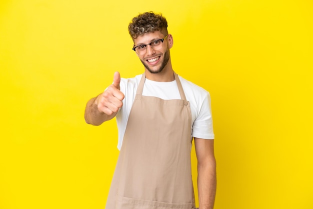 Camarero de restaurante hombre rubio aislado de fondo amarillo con los pulgares hacia arriba porque algo bueno ha sucedido