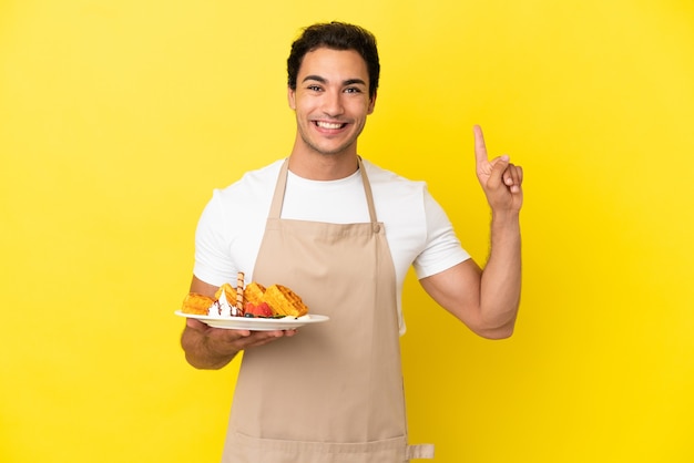 Camarero de restaurante sosteniendo gofres sobre fondo amarillo aislado apuntando hacia una gran idea