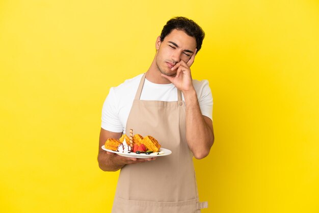 Camarero de restaurante sosteniendo gofres sobre fondo amarillo aislado con dolor de cabeza