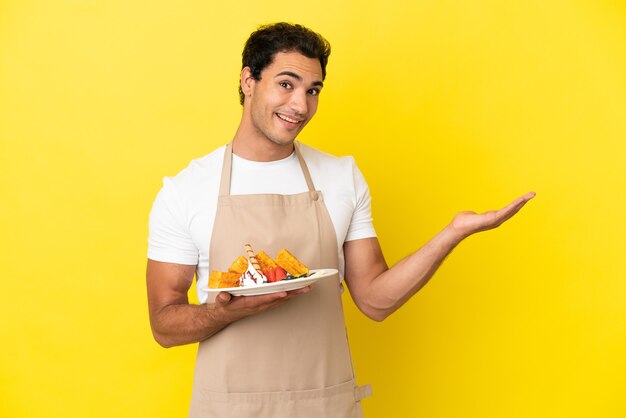 Foto camarero de restaurante sosteniendo gofres sobre fondo amarillo aislado extendiendo las manos hacia el lado para invitar a venir