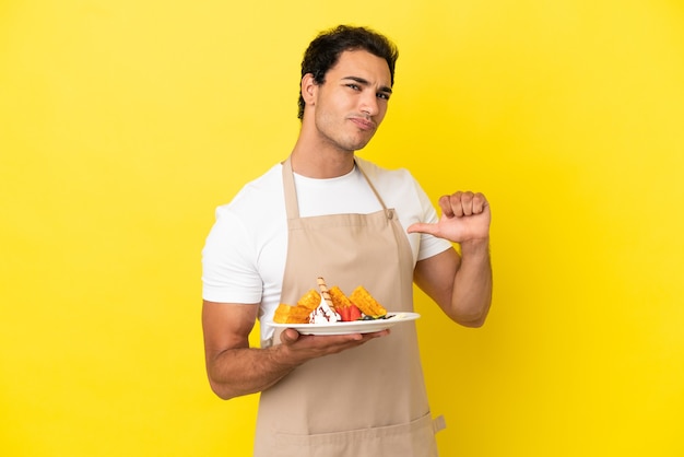 Camarero de restaurante sosteniendo gofres sobre fondo amarillo aislado orgulloso y satisfecho de sí mismo