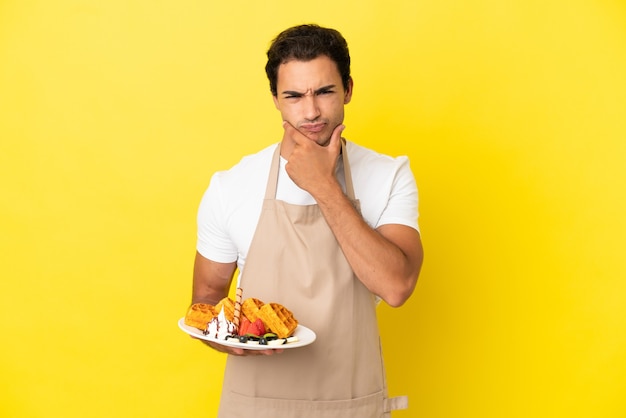 Camarero de restaurante sosteniendo gofres sobre fondo amarillo aislado pensando