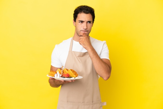 Camarero de restaurante sosteniendo gofres sobre fondo amarillo aislado teniendo dudas y pensando