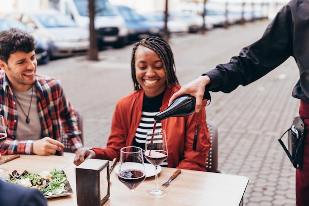Camarero sirviendo vino tinto a una mujer negra sonriente en la terraza de un bar