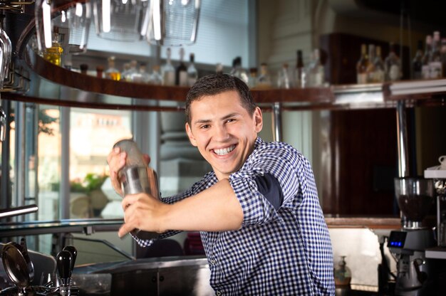 Camarero sonriente feliz detrás del mostrador de la barra agitando un cóctel en una coctelera de acero.