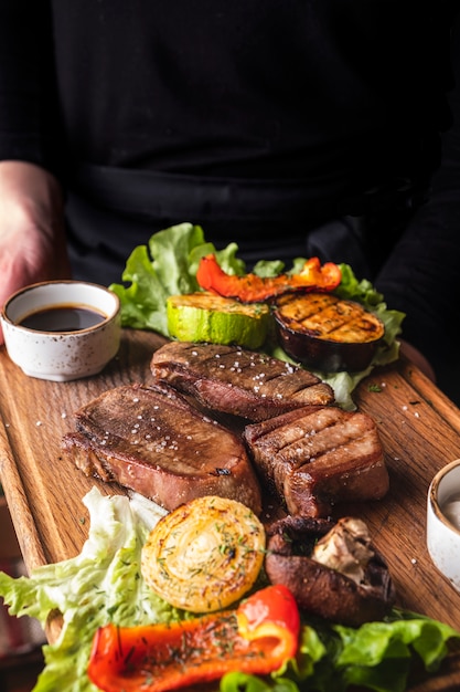 El camarero sostiene una tabla de madera con lengua de ternera a la parrilla y vegetales