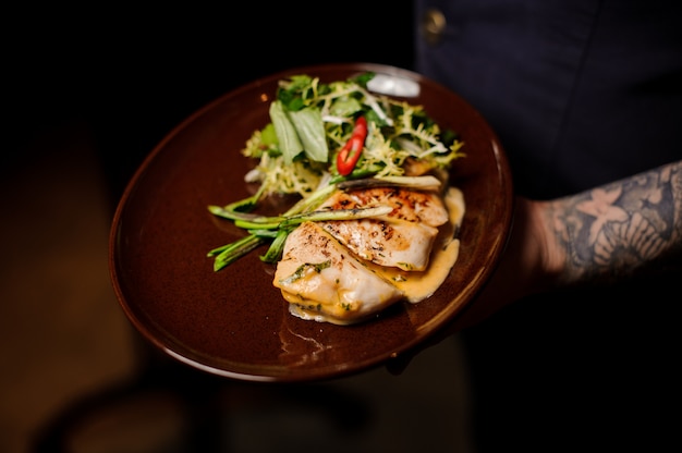 Camarero tatuado sosteniendo un plato de filete de pollo y ensalada