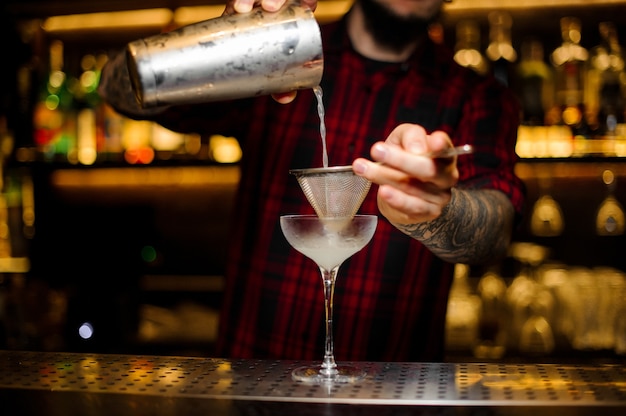 Camarero vertiendo bebida alcohólica fresca en un vaso elegante con un colador en la barra