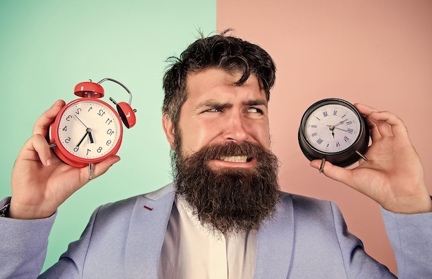 ¿Cambiar el reloj afecta tu salud? Un hombre hipster barbudo sostiene dos relojes diferentes. Un tipo sin afeitar con la cara perpleja que tiene problemas con el cambio de hora. Zona horaria. Los cambios de zona horaria afectan la salud.