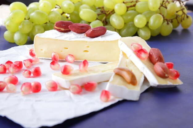 Camembert com sementes de romã, uvas verdes e nozes sobre fundo azul Delicioso queijo em papel pergaminho close-up