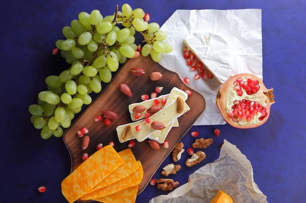 Camembert y lonchas de queso duro amarillo con semillas de granada y uvas verdes sobre un fondo azul Varios tipos de composición de queso en un primer plano de tablero de madera