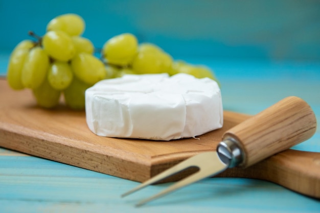 Camembert, mit einer Gabel für Käse und Trauben auf einem Schneidebrett, auf blauem Holzhintergrund