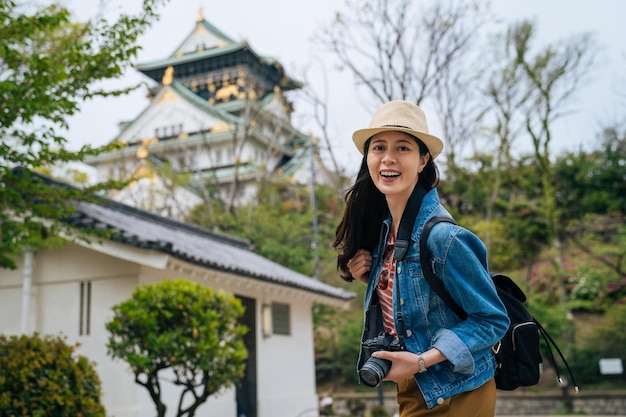 câmera de rosto jovem turista sorrindo segurando a câmera slr. fotógrafa profissional sempre viaja com sua digicam. senhora viajante visita o castelo himeji de osaka em dia ensolarado com árvores verdes ao redor.