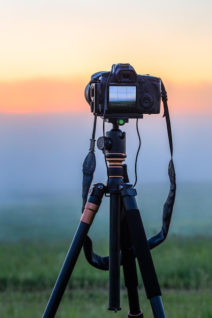 Câmera digital preta em tripé fotografando paisagem matinal nebulosa no verão com foco seletivo