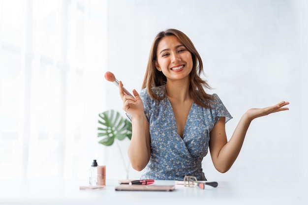 Câmera olhando Jovem vlogger de beleza asiática fazendo um tutorial de maquiagem ao sentar na mesa na sala de estar