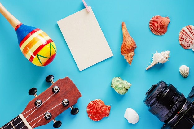 Câmera, ukulele, maracas, cartão postal de papel em branco e conchas do mar em fundo azul