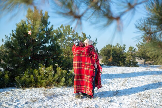 Caminando en el invierno en el bosque, un chico envuelve a su novia en una cálida tela escocesa roja para que se caliente