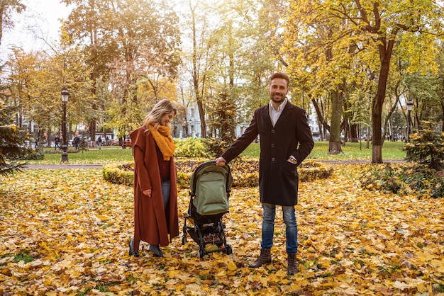 Caminando en un parque de otoño familia joven con un bebé recién nacido en un cochecito