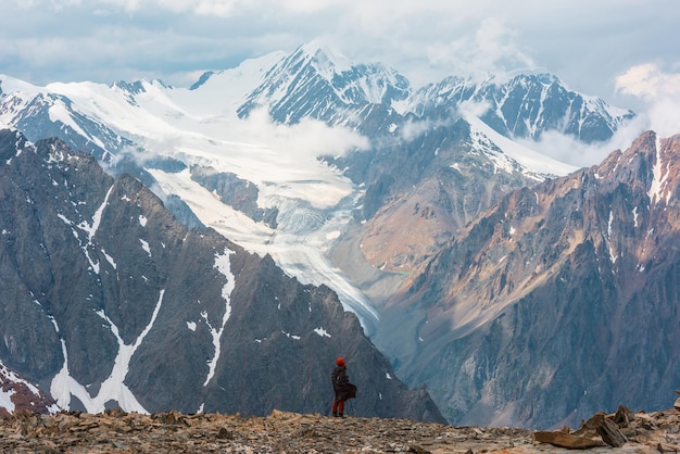 Caminante a gran altura con vistas a la larga lengua glaciar y altas montañas nevadas bajo un cielo nublado Hombre en una colina de piedra ventosa contra enormes montañas con nubes bajas Hombre y majestuosas montañas naturaleza