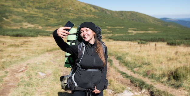 Caminante de mujer se encuentra en la cima de la montaña, sostiene un teléfono en sus manos y toma una selfie en su teléfono inteligente