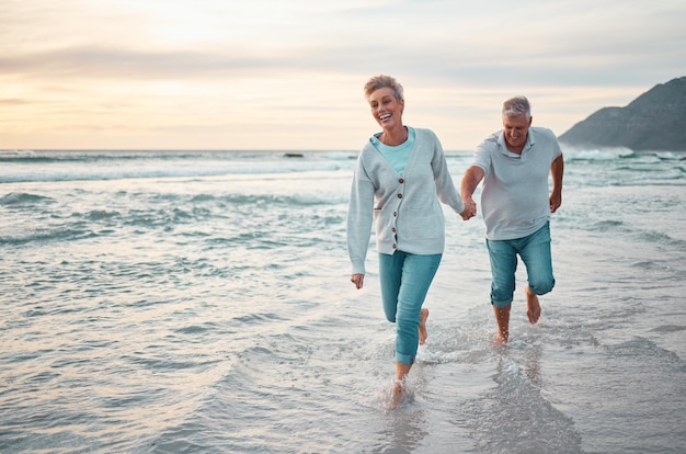 Caminar por la playa y una pareja mayor tomándose de la mano para apoyar el amor y el cuidado con la jubilación de bienestar al aire libre y el estilo de vida de vacaciones con el cielo del atardecer Personas mayores corriendo juntas en el agua del océano