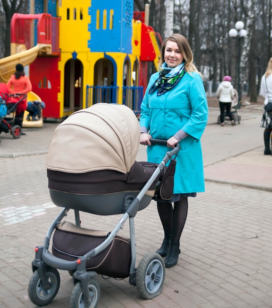 Caminhada na cidade com carrinho jovem mãe ativa em casaco azul andando no playground com seu bebê no carrinho