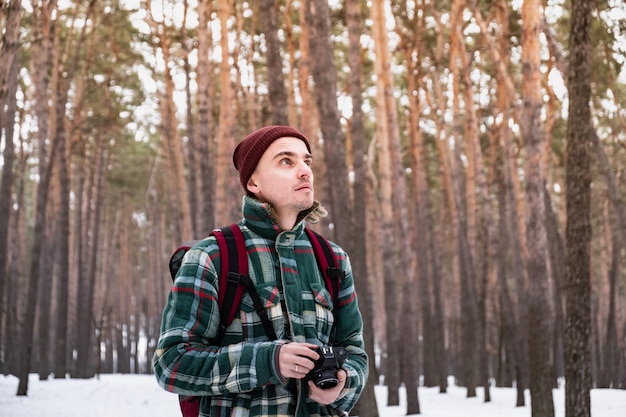 Caminhadas pessoa masculina na floresta de inverno tirando fotografias. Homem de camisa quadriculada de inverno no belo bosque nevado com uma câmera de filme antigo