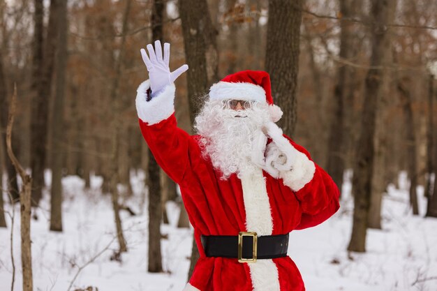 Caminhando pela floresta de inverno, o papai noel acena com a mão carregando presentes de natal