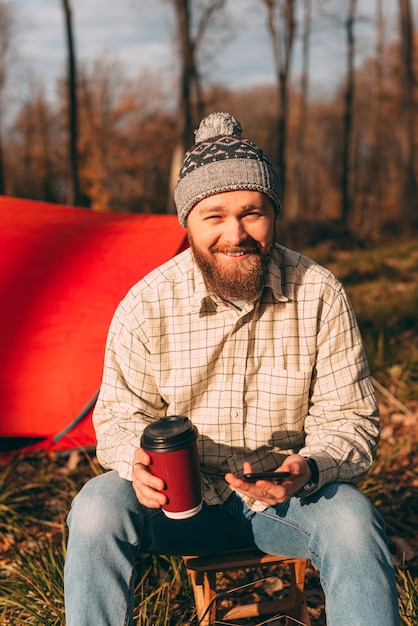 Caminhante barbudo feliz com smartphone e xícara na floresta