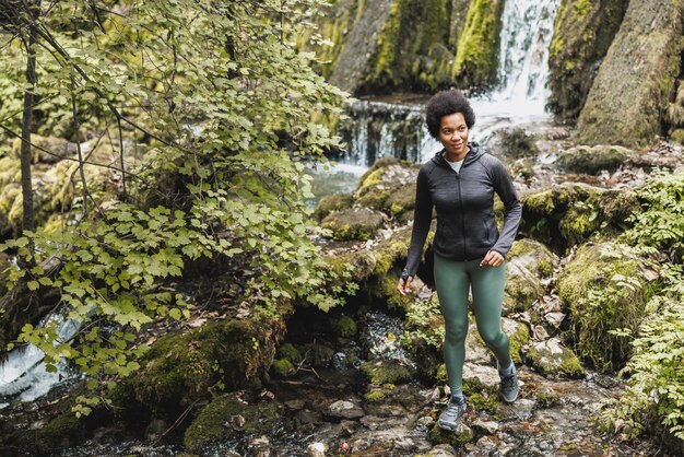 Caminhante de mulher afro-americana madura atravessando um riacho perto da cachoeira enquanto explorava as montanhas e desfrutava de treinamento ao ar livre.