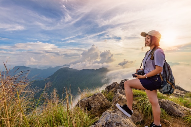 Caminhante Menina adolescente fofa asiática feliz com a mochila da câmera e boné olhando a bela paisagem do céu da natureza ao pôr do sol na montanha, Viagem de aventura na Ásia no Parque Florestal de Phu Chi Fa, Chiang Rai, Tailândia