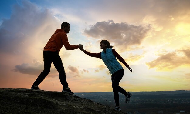 Caminhantes de homem e mulher, ajudando uns aos outros a escalar a pedra ao pôr do sol nas montanhas.